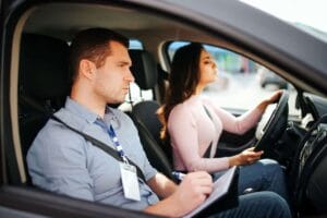 Egzaminator siedzi na miejscu pasażera i robi notatki, podczas gdy kobieta prowadzi samochód w trakcie egzaminu na prawo jazdy.