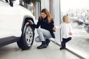 Kobieta z małym dzieckiem ogląda samochód w salonie – sprawdza oponę stojącego pojazdu.