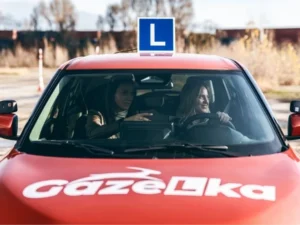 Kursantka prowadzi samochód nauki jazdy GAZeLka z oznaczeniem „L”, a instruktor udziela wskazówek podczas jazdy szkoleniowej.